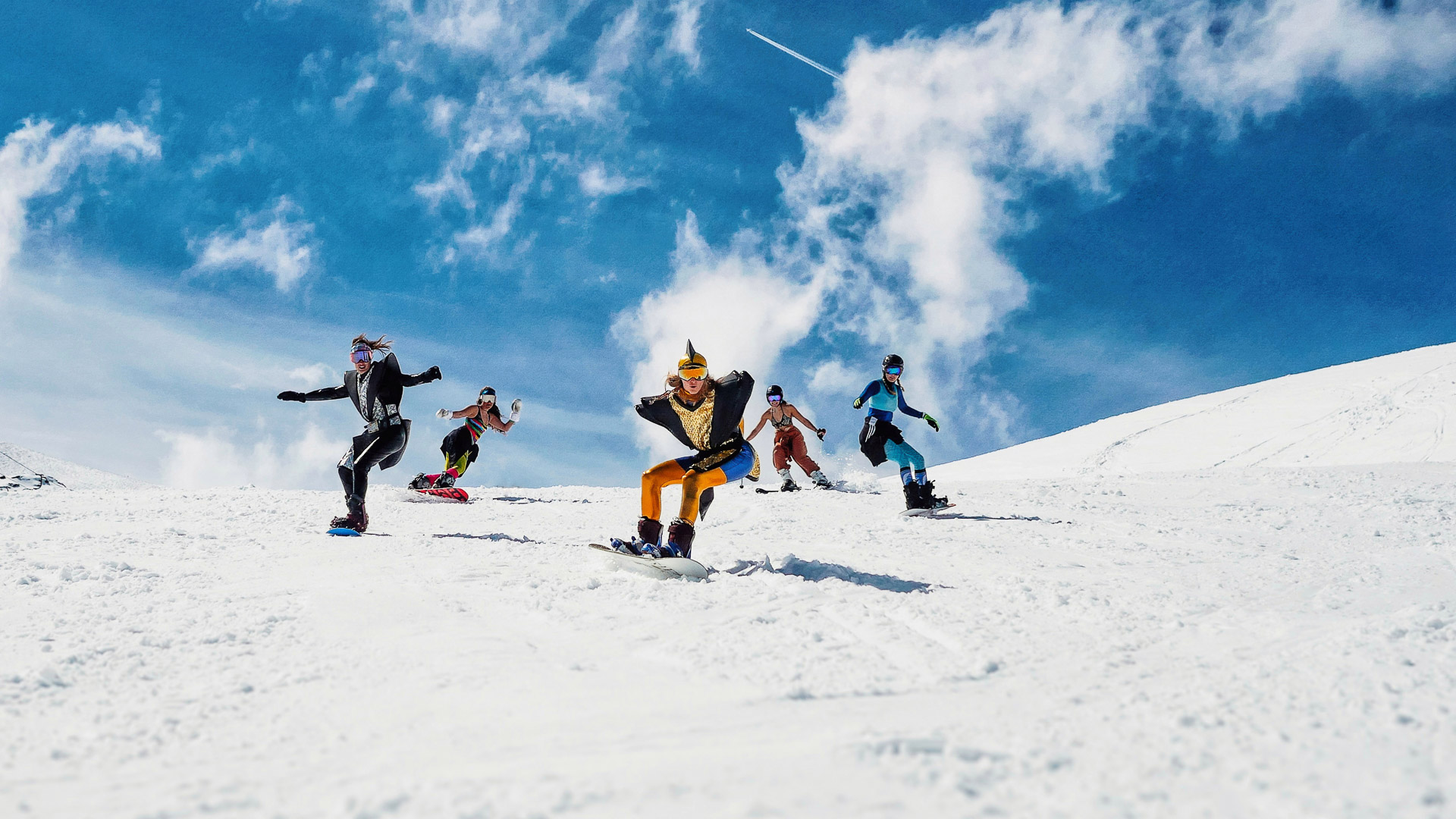 Snowboarderinnen und Skifahrerinnen fahren in lustigen Outfits die Piste herunter.