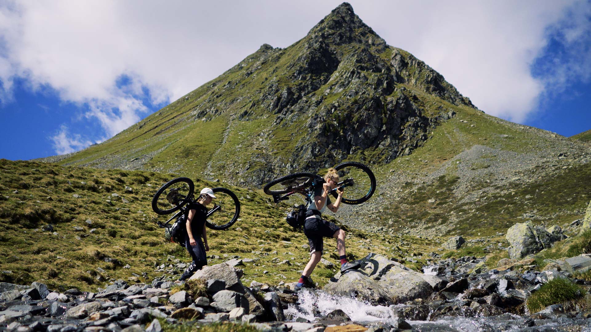 Two women carry their mountain bikes across a river in the Alps, a steep mountain is in the background.